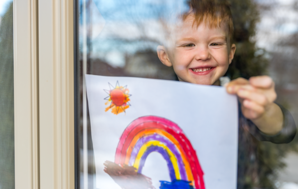 A kid holding up a painted rainbow in the window.
