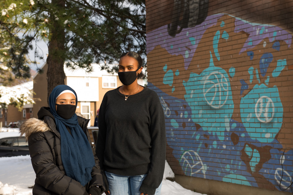A photo of Noor standing outside next to a brick wall with grafitti.
