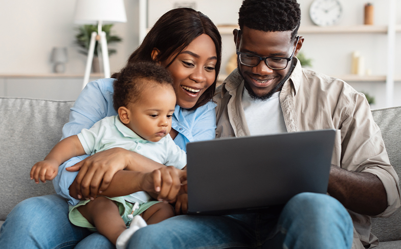 Smiling family sitting on a couch, looking at a laptop together. A mother holds a baby on her lap while the father navigates the computer.
